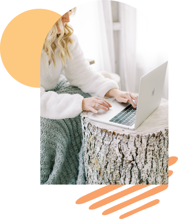 Women typing on a laptop sitting at a rough log table with a knit blanket and cozy sweater. 