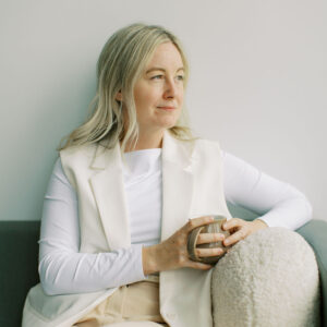 A person with long hair, a registered social worker, holds a mug while sitting on a gray couch at The Cove. They wear a white outfit and look thoughtfully into the distance, as if reflecting on their next therapy session.