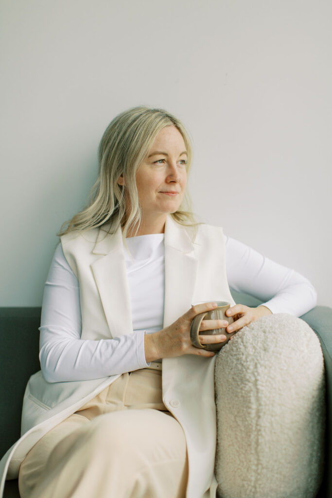 A person with long hair, a registered social worker, holds a mug while sitting on a gray couch at The Cove. They wear a white outfit and look thoughtfully into the distance, as if reflecting on their next therapy session.