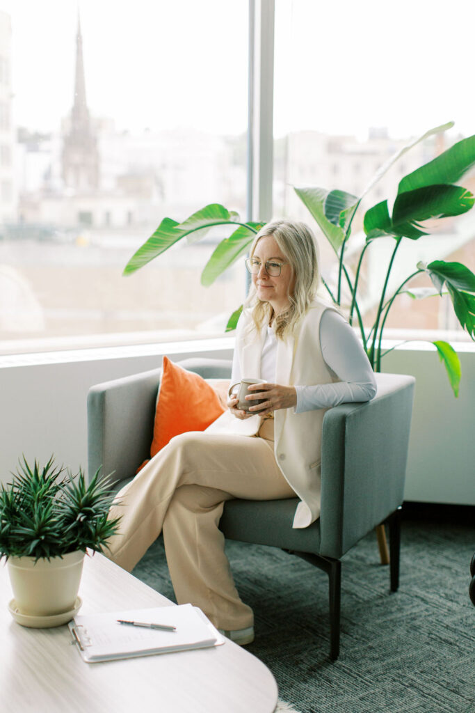 A person sits in a chair by the window at The Cove, holding a mug. A thriving plant adds tranquility nearby, while a notepad on the table invites reflection—an ideal setting for nurturing mental health through mindful therapy and introspection.