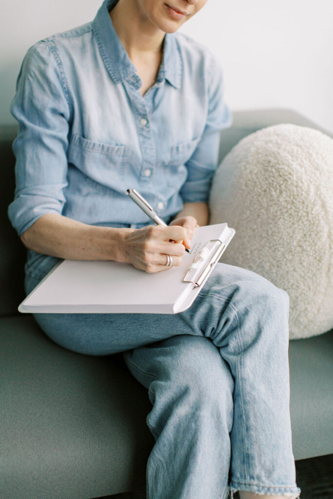 A person in denim clothing sits on a gray sofa at The Cove, meticulously writing on a clipboard, embodying the calm focus of a therapy session dedicated to mental health exploration.