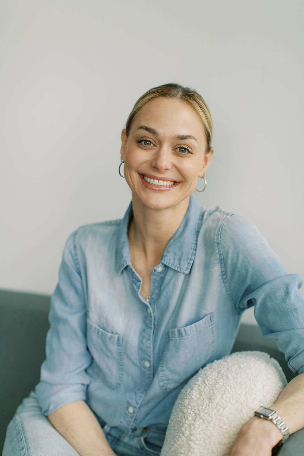 A person with a denim shirt smiles while seated on a sofa, embodying the warmth and compassion of a registered psychotherapist deeply committed to promoting mental health.