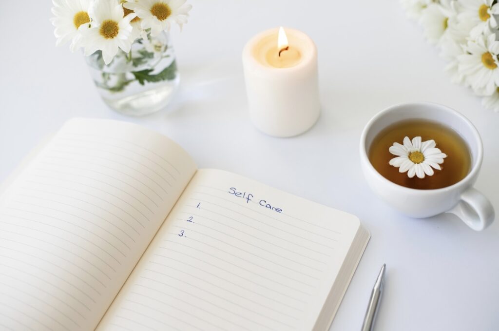 A notebook with "Self Care" written on a page, a lit candle, a cup of tea with a daisy, and a vase of white flowers on a white surface