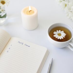 A notebook with "Self Care" written on a page, a lit candle, a cup of tea with a daisy, and a vase of white flowers on a white surface