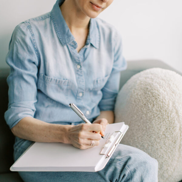 Person in light denim clothing seated on a gray couch, writing on a clipboard resting on their lap.