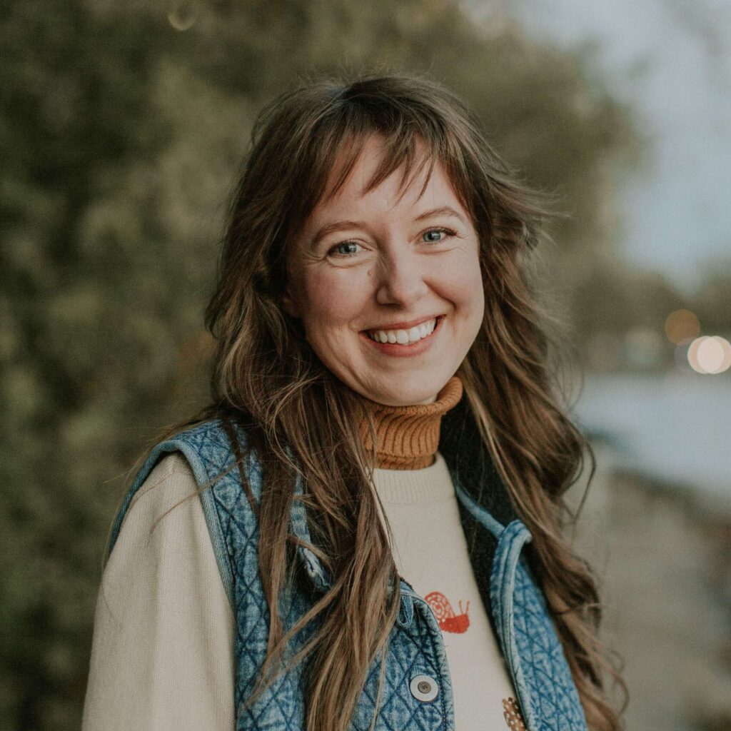 A woman with long hair smiles outdoors, wearing a blue quilted vest over a beige sweater with colorful patterns. A blurred natural background is visible.