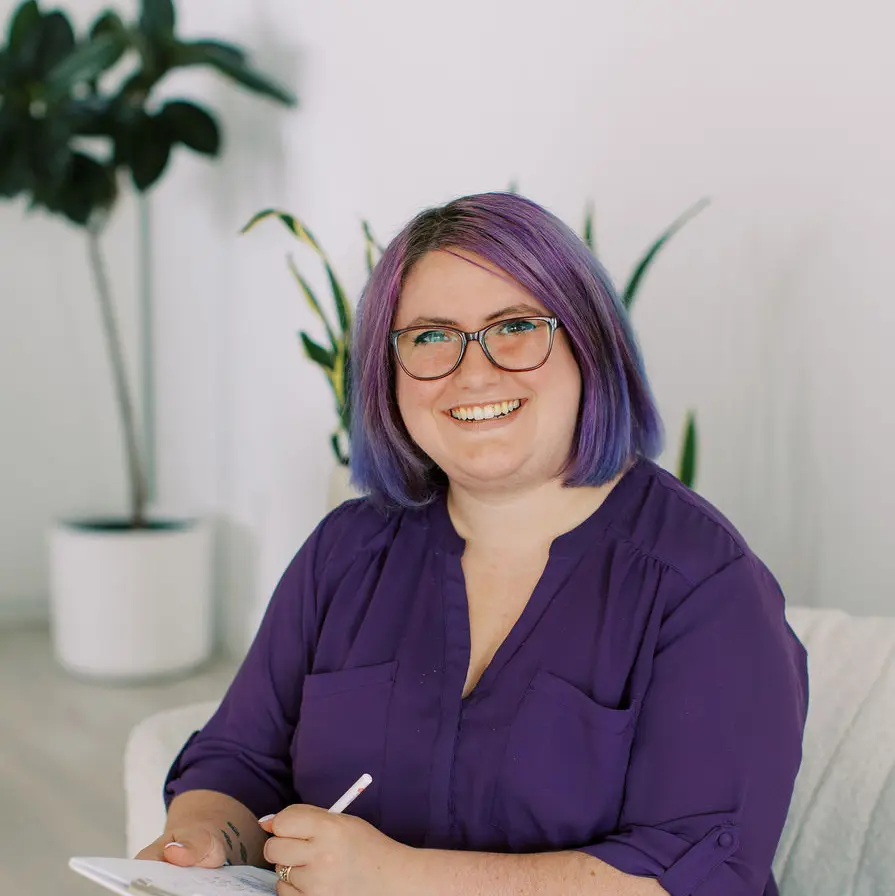 A therapist with purple hair and glasses sits on a couch, smiling and writing in a notebook. A potted plant is in the background.
