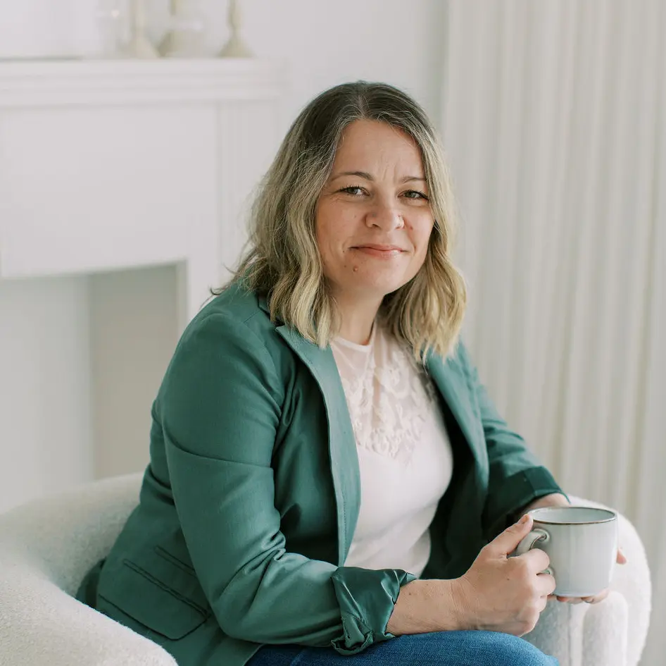 Therapist with shoulder-length blonde hair sits in a chair, holding a mug, wearing a green blazer and white blouse, in a softly lit room.
