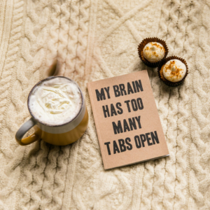 A mug of frothy drink, two cupcakes, and a notebook with “MY BRAIN HAS TOO MANY TABS OPEN” on a beige knit blanket.