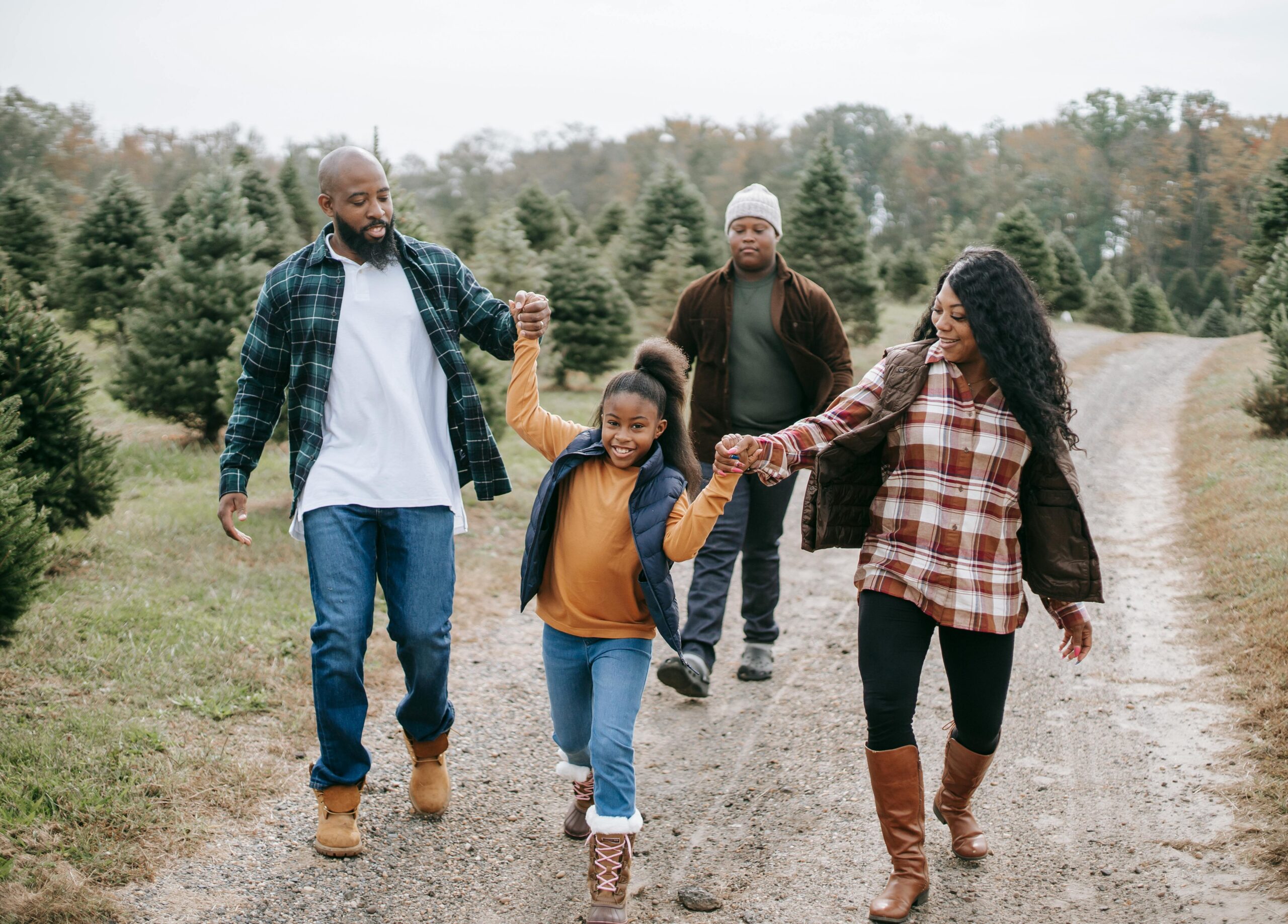 A family of four holding hands and walking together down a path at a Christmas tree farm, smiling.